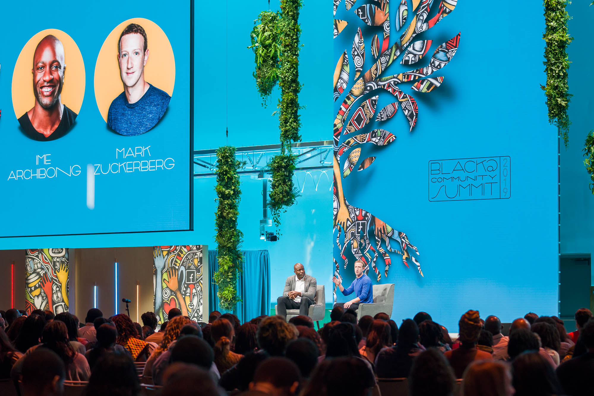 Ime Archibong and Mark Zuckerberg seated on stage speaking at the 2019 Black Community Summit with an audience in front and colorful tree artwork on a blue background.