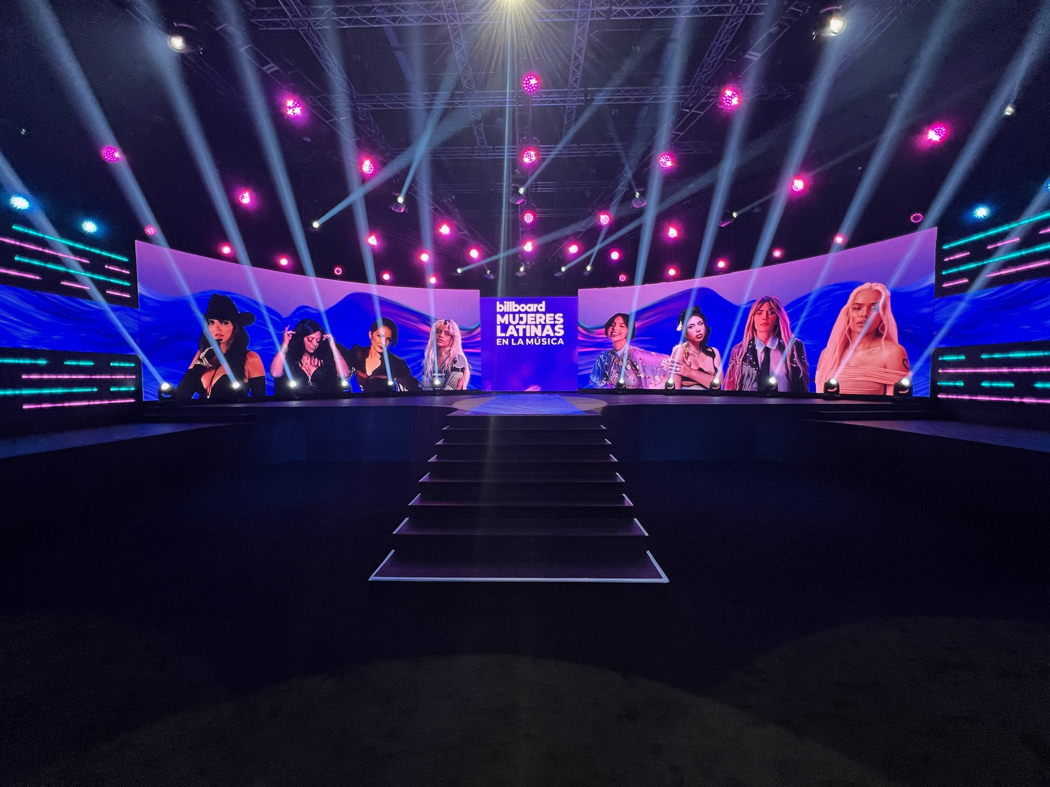 Stage with large screens displaying images of Latin female musicians and the text 'billboard MUJERES LATINAS EN LA MÚSICA' illuminated by colorful lights and spotlights.