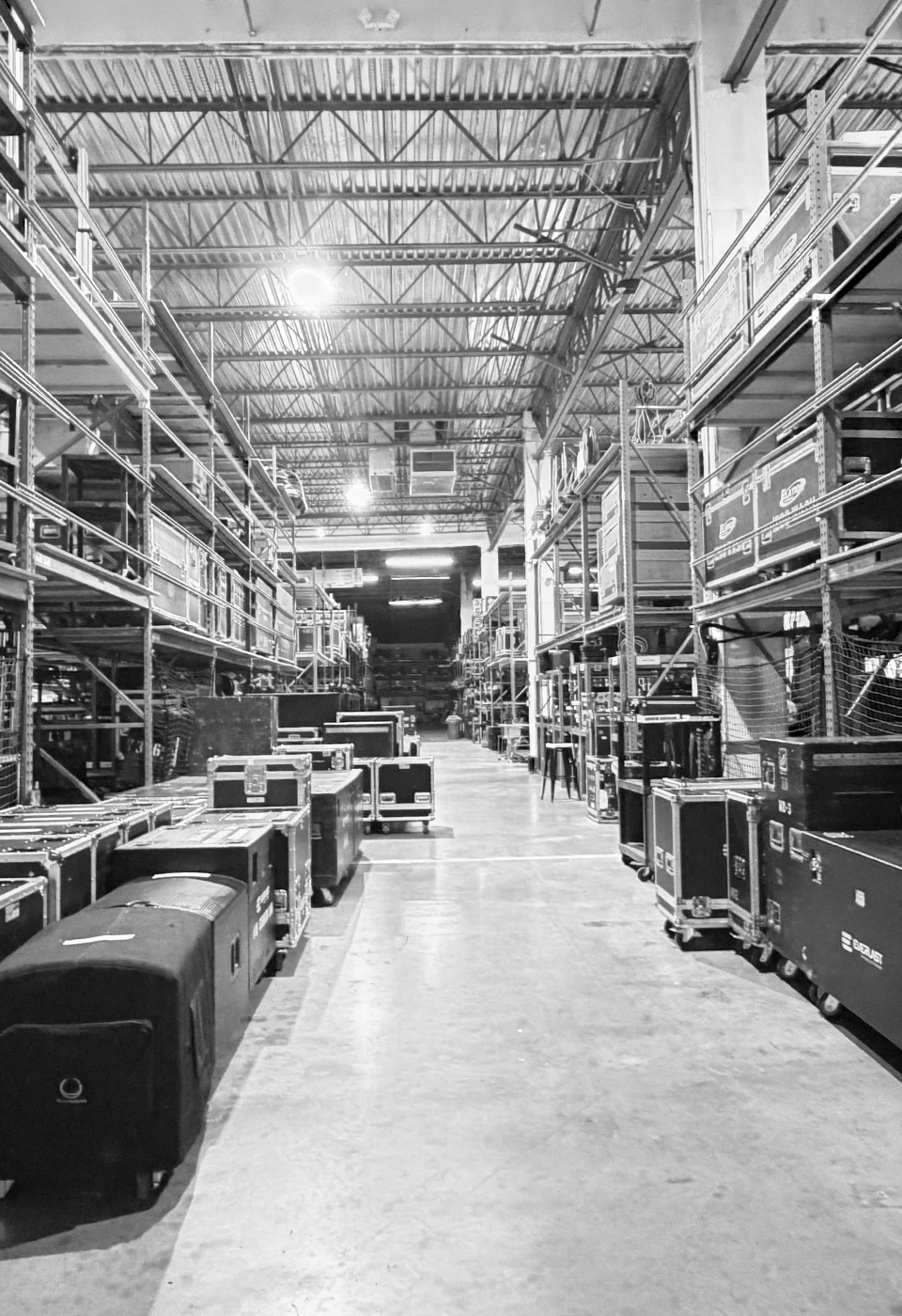 Interior of a large warehouse with metal shelves filled with equipment cases and storage boxes along a wide aisle.