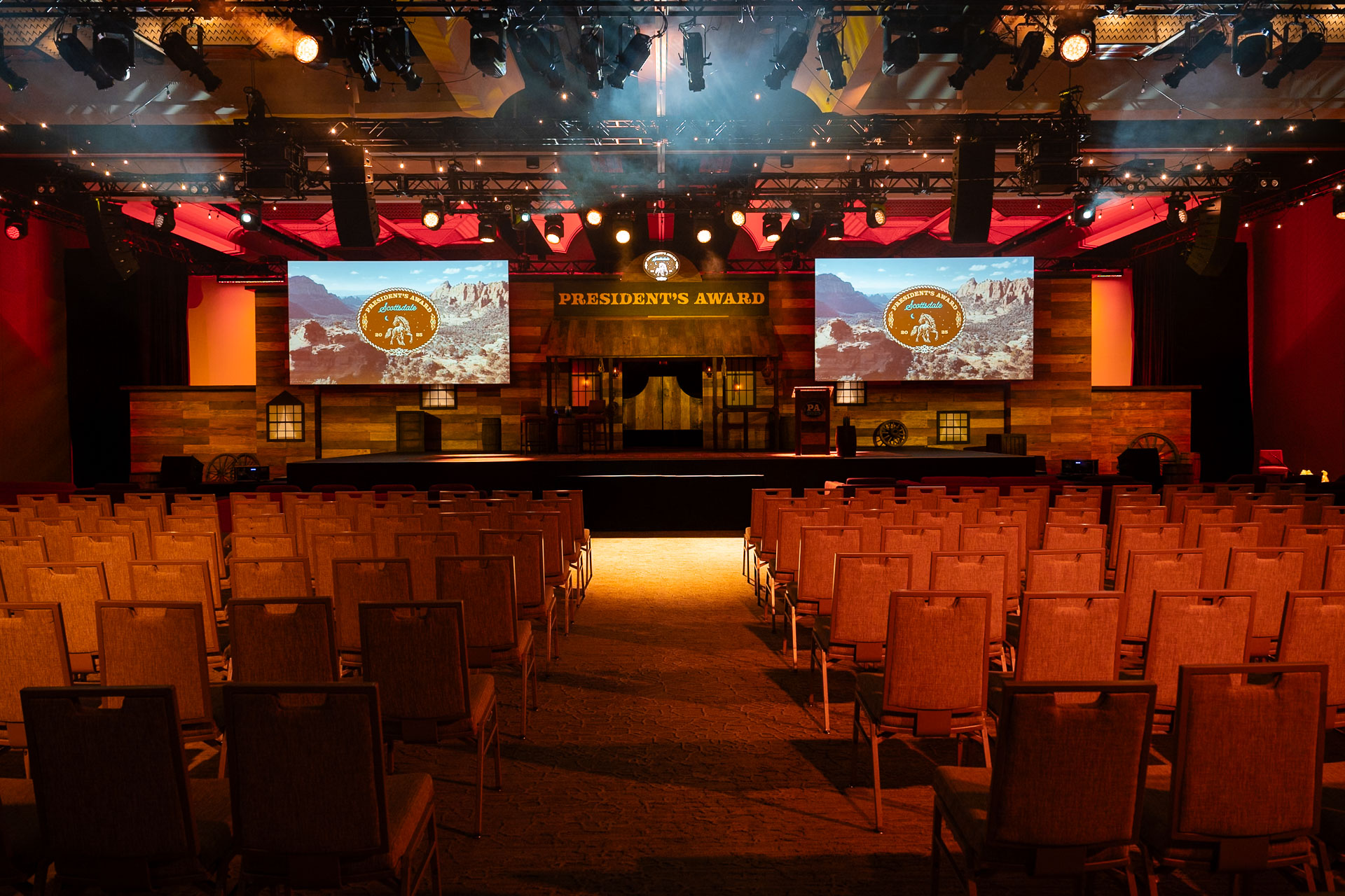 Empty auditorium with rows of chairs facing a stage displaying 'President's Award' with dual large screens showing a desert landscape and badge.