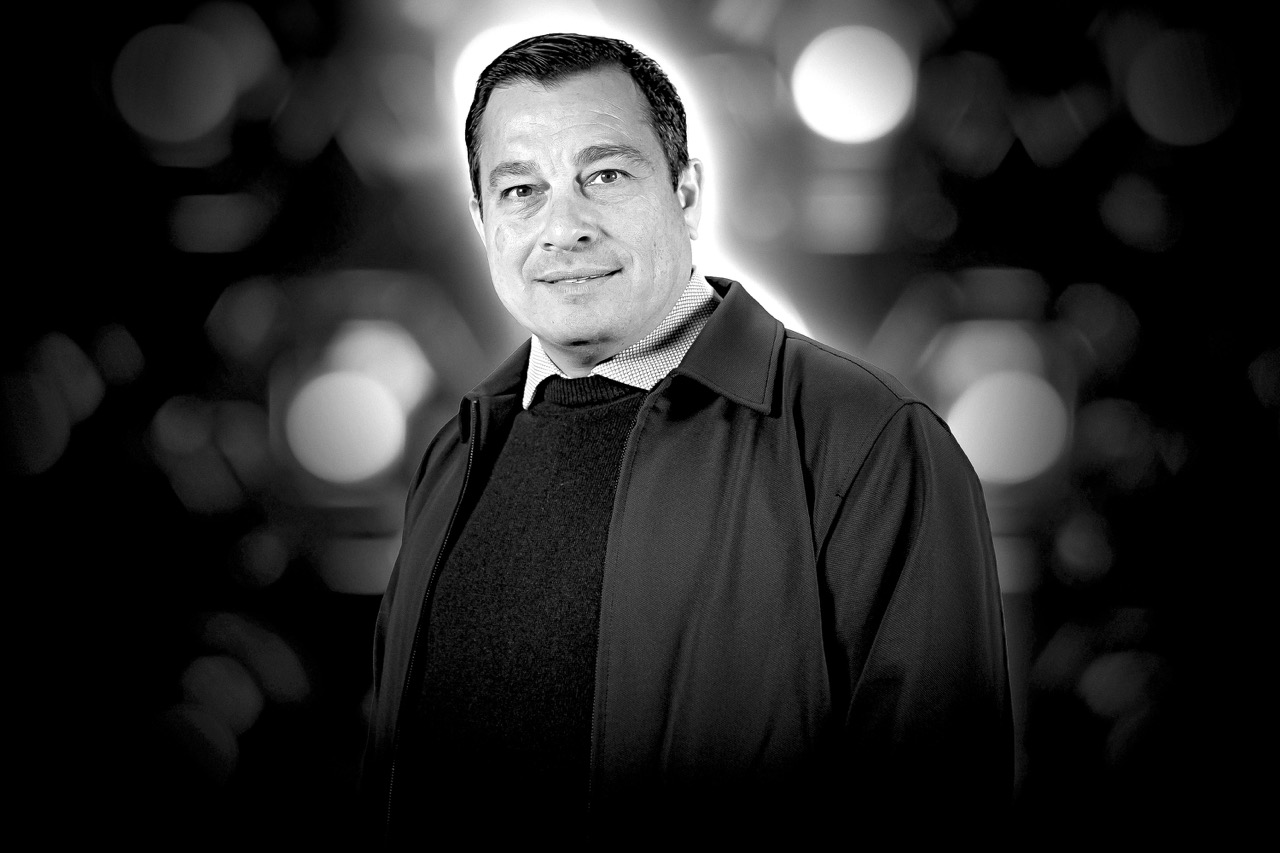 Black and white portrait of a man wearing a jacket and sweater, smiling slightly against a blurred background with circular light bokeh.