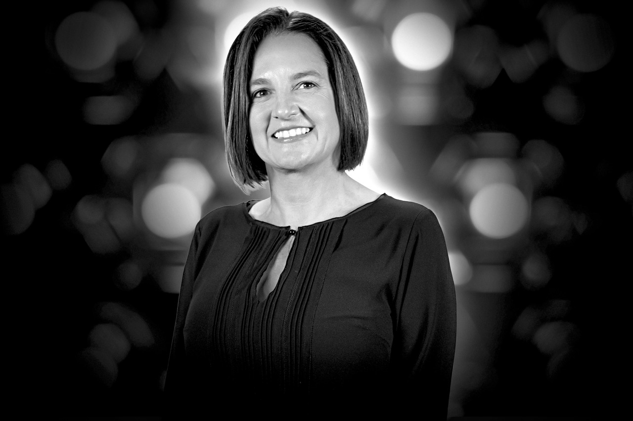 Smiling woman with straight shoulder-length hair wearing a dark top against a blurred background with circular light patterns.