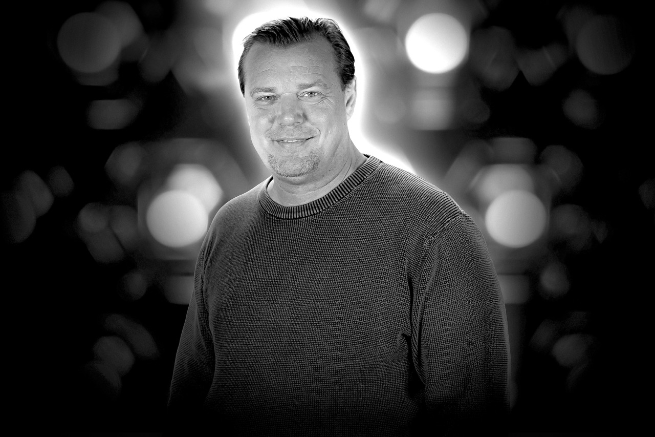 Middle-aged man with short hair wearing a knit sweater, smiling against a blurred dark background with bright bokeh lights.