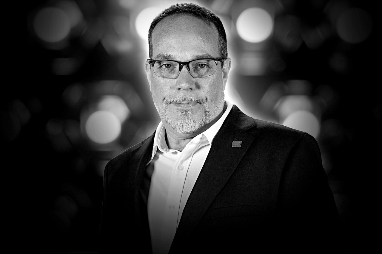 Middle-aged man with glasses and short beard wearing a dark blazer and white shirt against a blurred background.