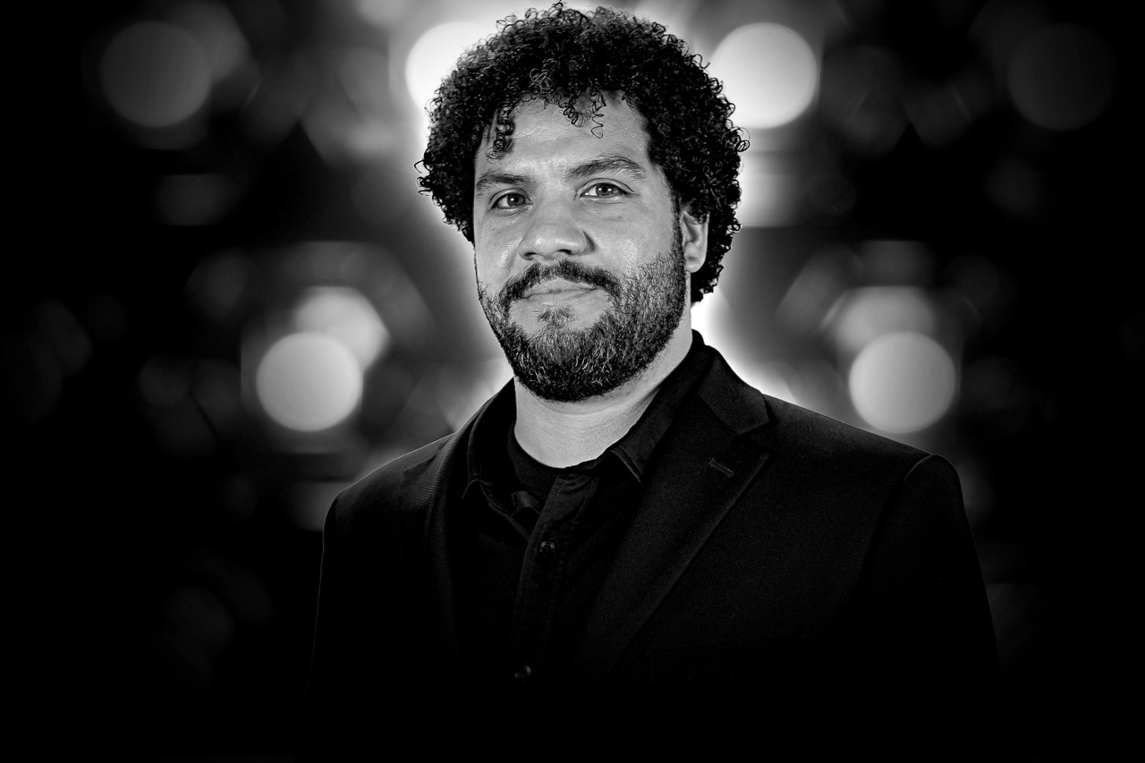 Black and white headshot of a man with curly hair and beard wearing a dark blazer and shirt, with blurred lights in the background.