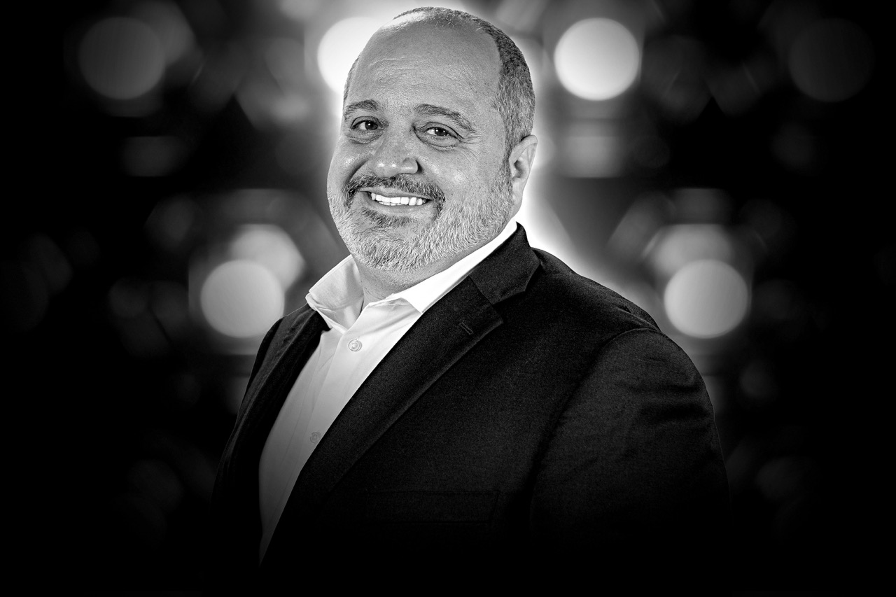 Smiling middle-aged man with a trimmed beard wearing a dark suit jacket and white shirt, against a blurred light background.