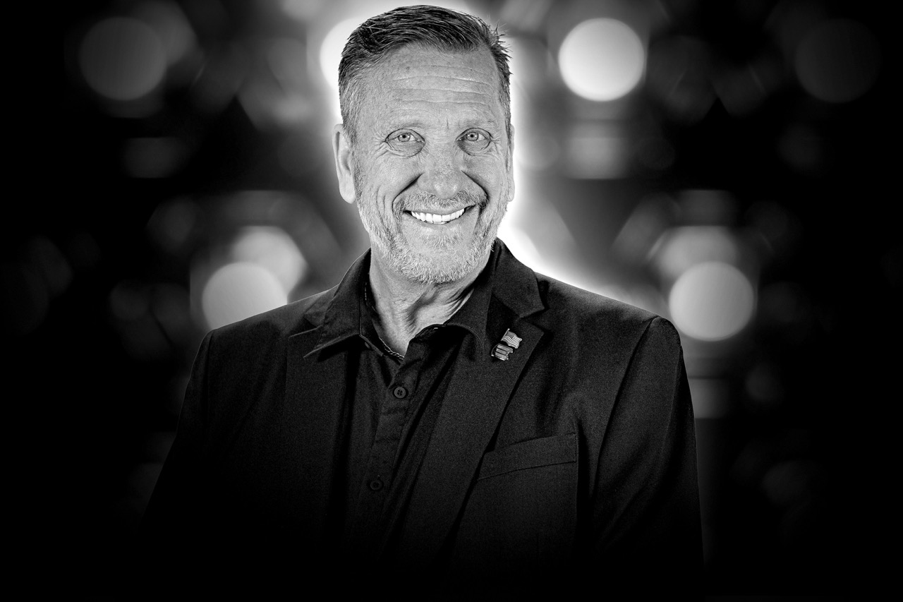 Smiling middle-aged man in a dark blazer with a lapel pin against a blurred background.