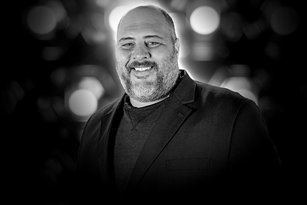 Smiling bearded man wearing a dark blazer and shirt with blurred circular lights in the background.