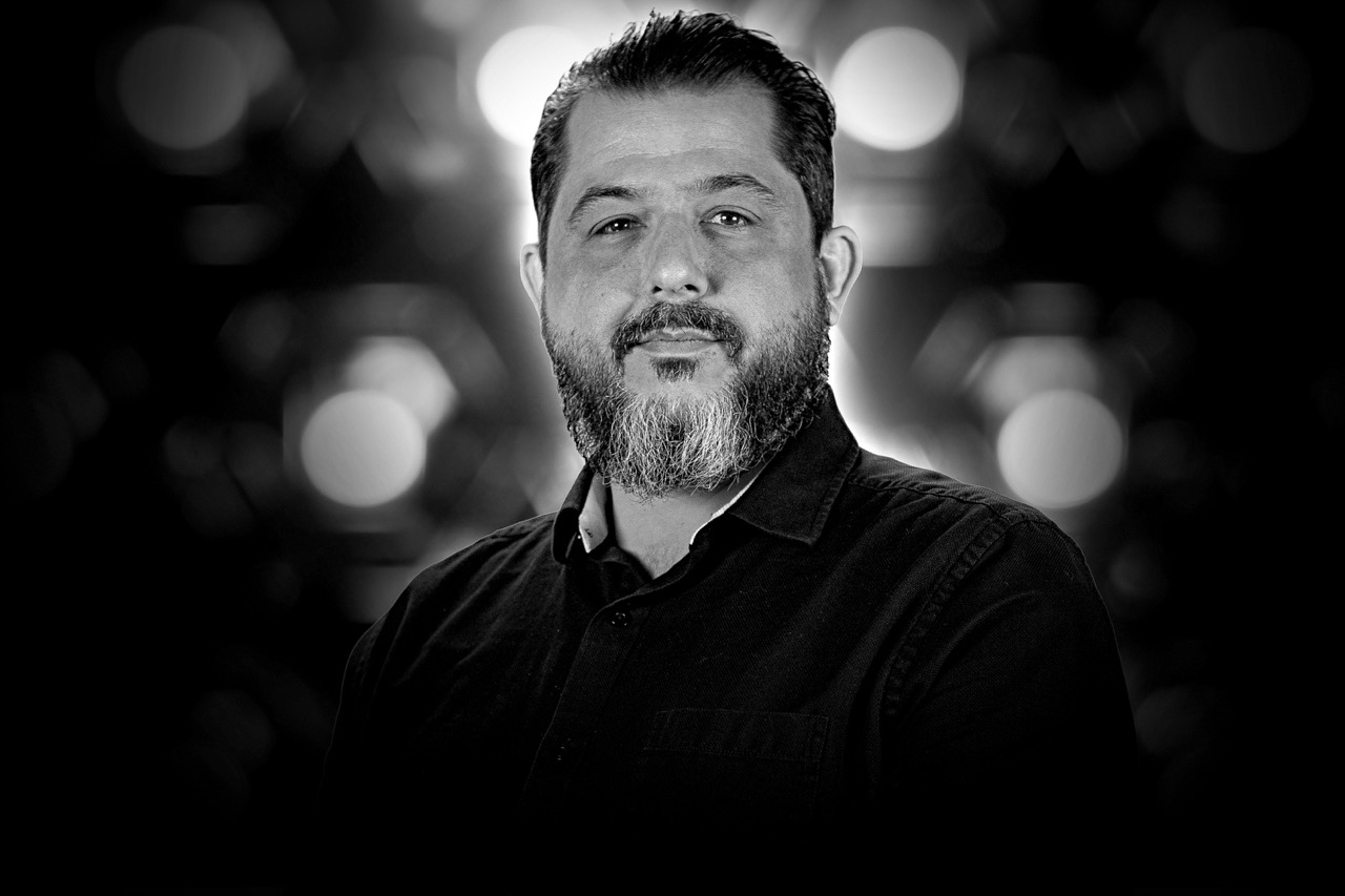 Black and white portrait of a man with a dark beard and shirt, with blurred circular lights in the background.