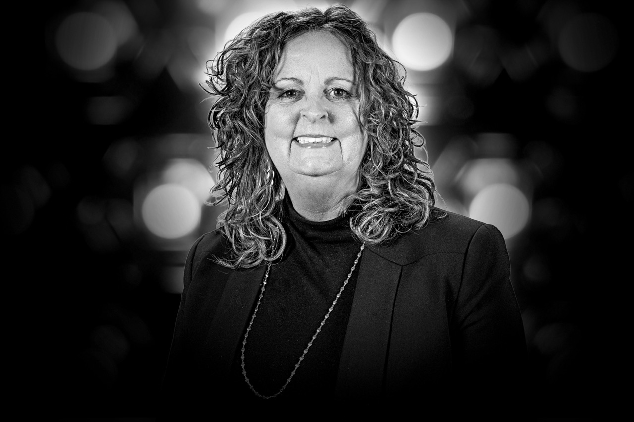 Black-and-white portrait of a smiling woman with curly hair wearing a dark blazer and a long necklace.