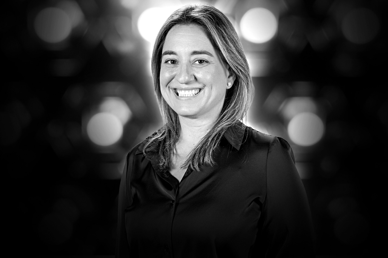 Smiling woman with shoulder-length hair wearing a black blouse against a dark, blurred background with bright circular lights.