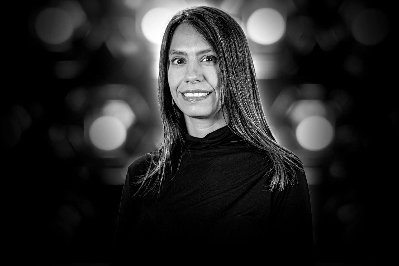 Black and white portrait of a smiling woman with straight hair wearing a black turtleneck against a background with blurred circular lights.