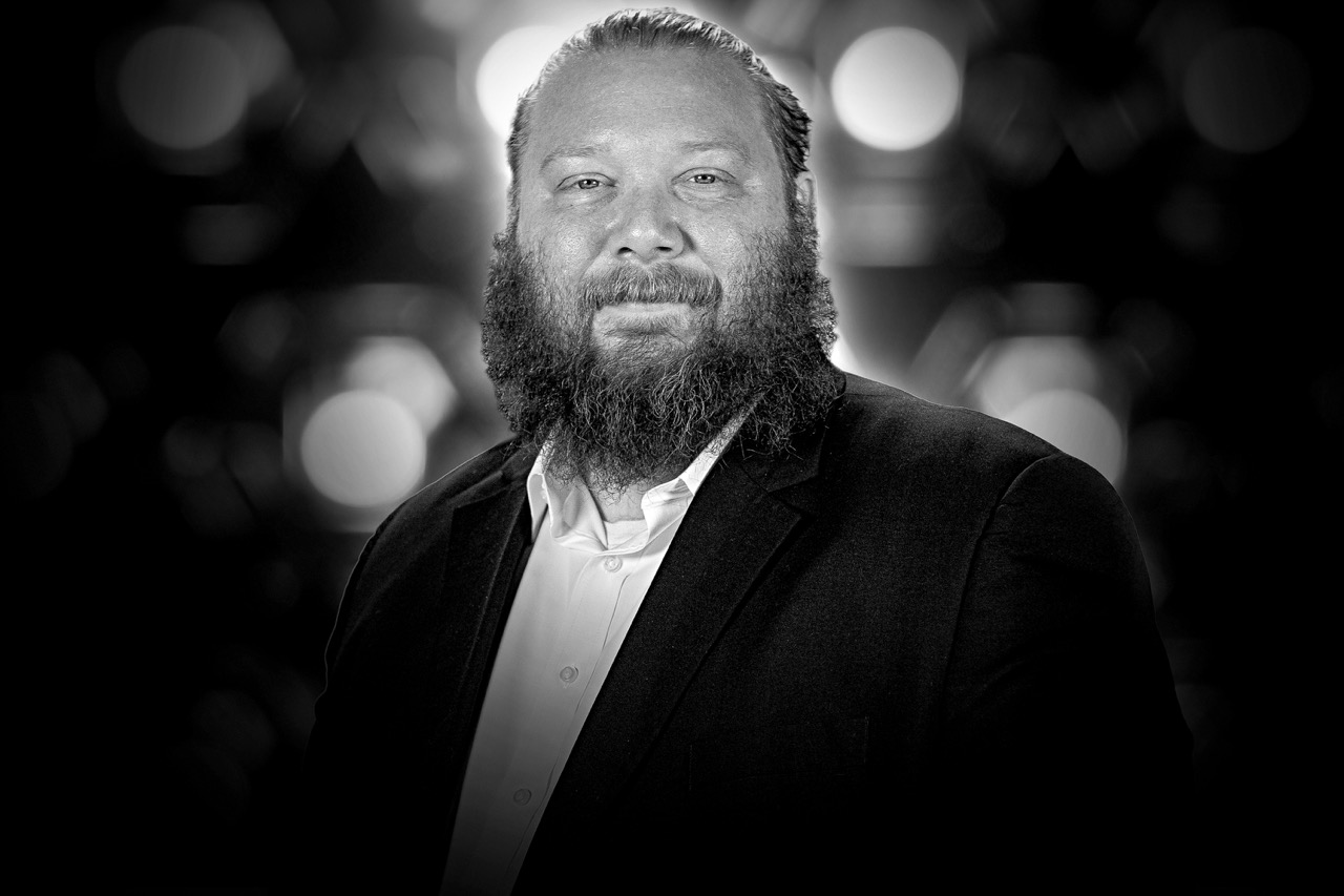 Portrait of a bearded man in a black suit jacket and white shirt against a blurred background with bokeh lights.