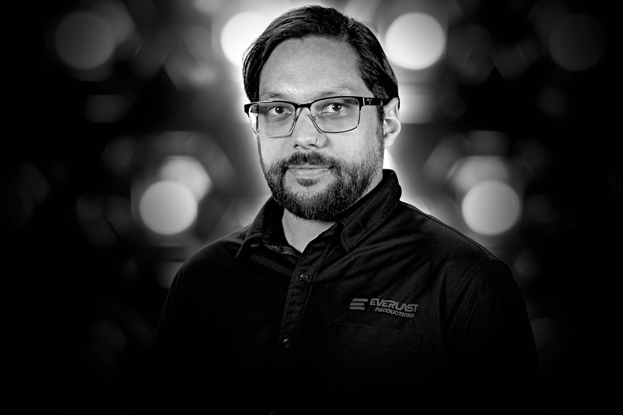 Black and white headshot of a bearded man with glasses wearing a shirt with Everlast Productions logo against a blurred background.