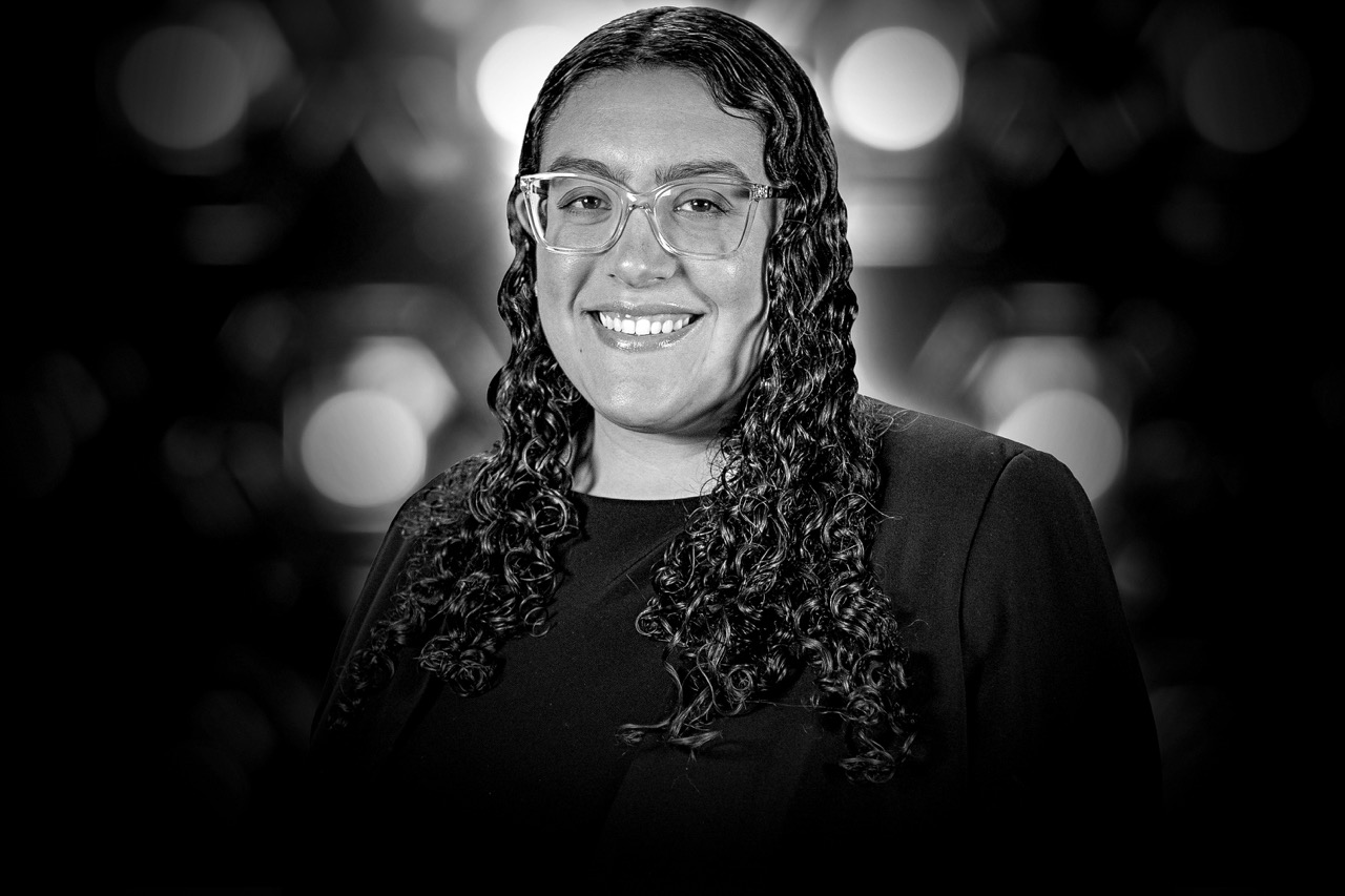 Black and white headshot of a person with long curly hair, clear glasses, and a dark blazer smiling against a blurred background.