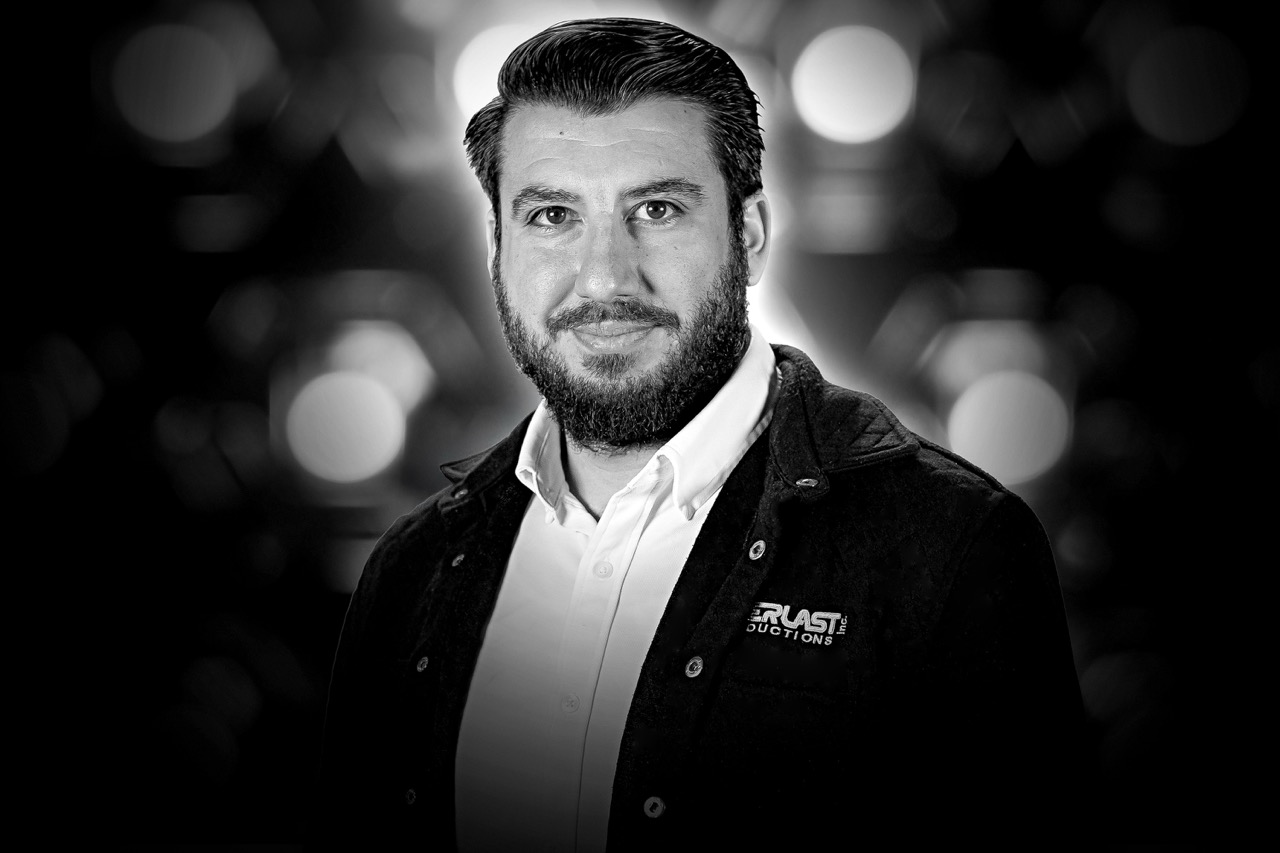 Man with dark hair and beard wearing a white shirt and dark jacket against a blurred background with circular light patterns.