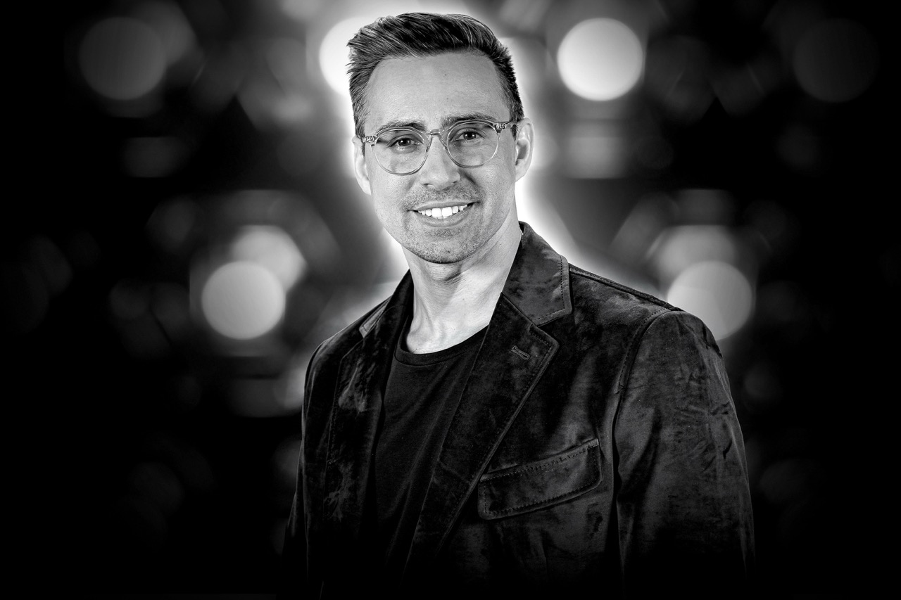 Smiling man with glasses wearing a dark blazer and t-shirt against a blurred background with circular lights.