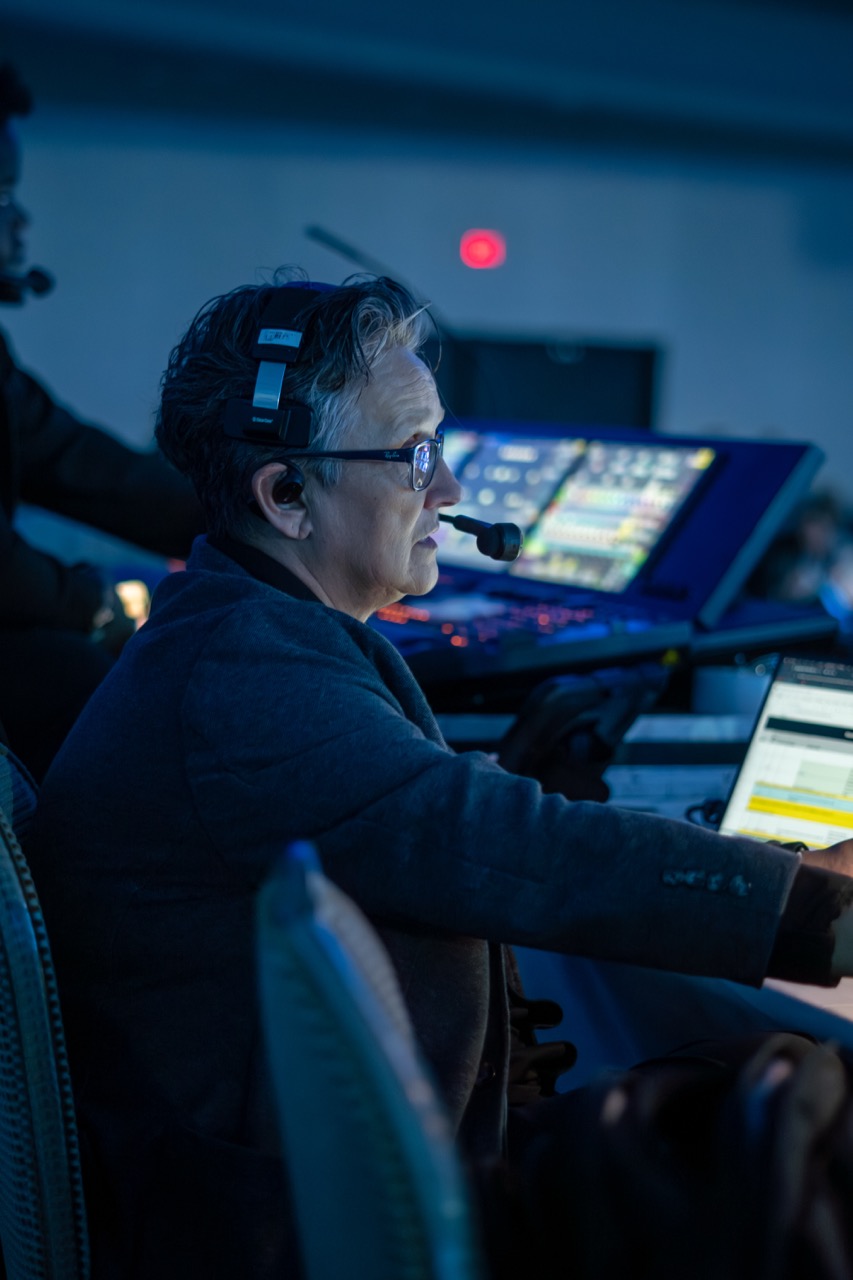 Person wearing headset and glasses working at a control panel with illuminated screens in a dimly lit room.