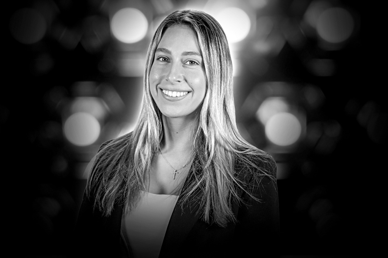 Smiling woman with long light hair wearing a blazer and a cross necklace against a blurred light background.