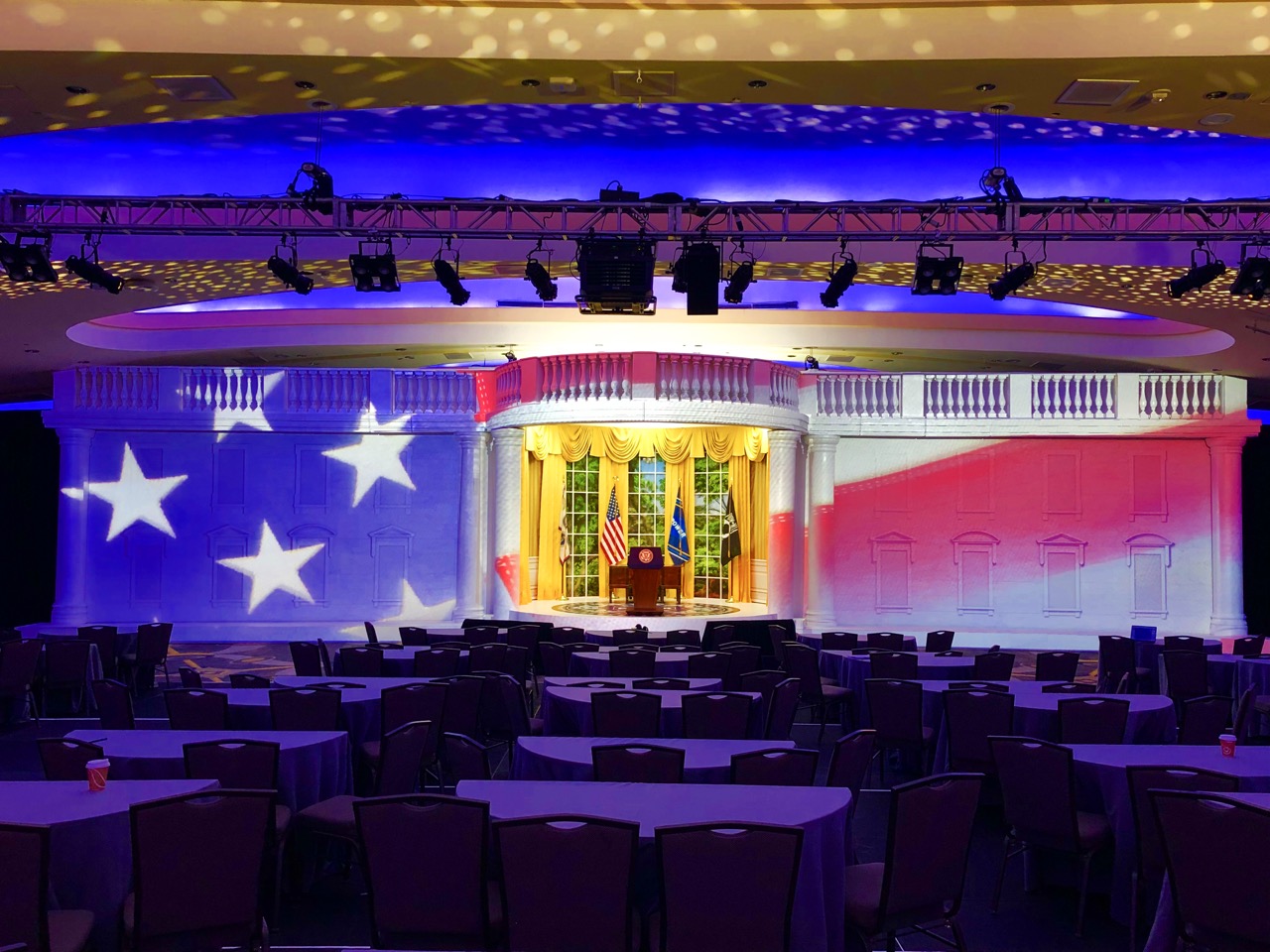 Indoor event space with a stage backdrop resembling the White House Oval Office illuminated with an American flag pattern and rows of empty round tables and chairs in front.