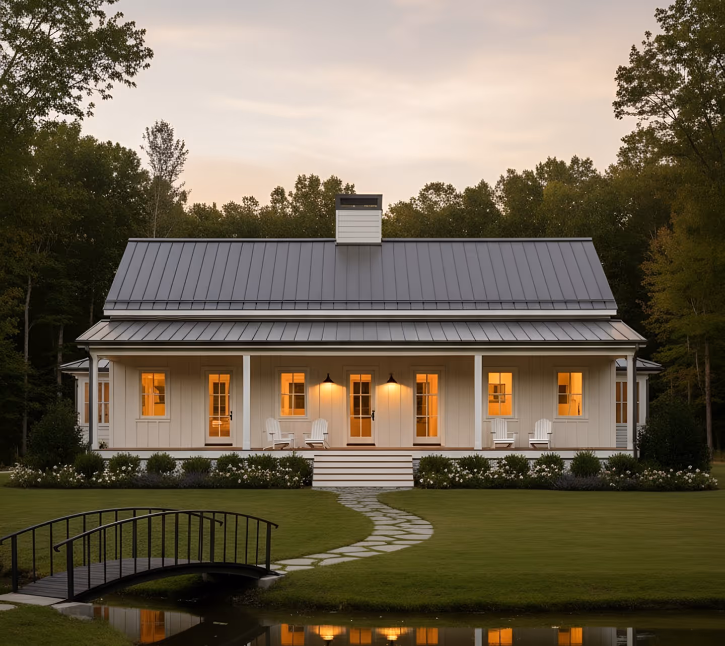 Modern white house with metal roof, lit windows, porch with chairs, stone path, small arched bridge, and reflecting pond in front.