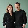 Smiling man and woman standing side by side against a green background, both wearing black tops.