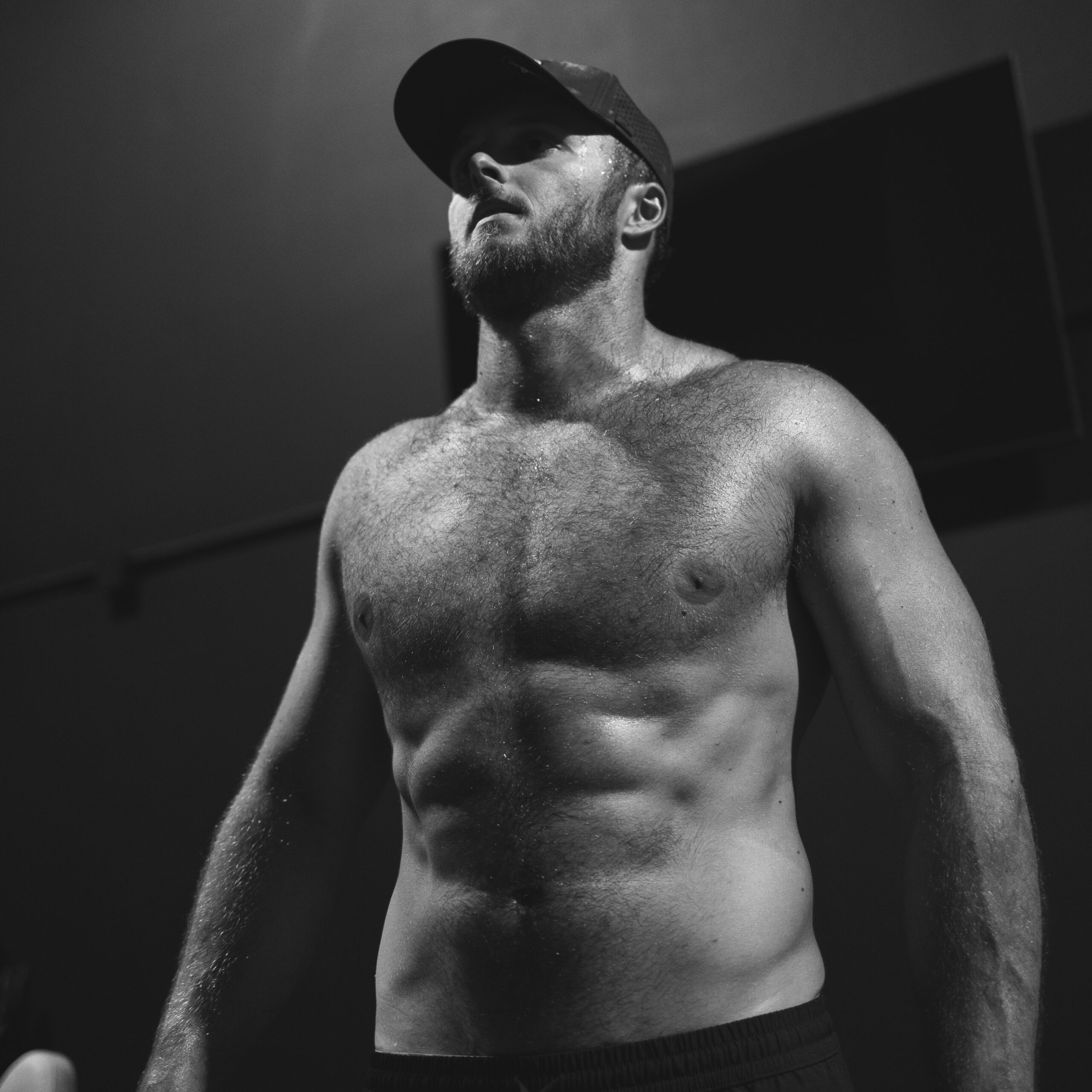 Black and white photo of a muscular shirtless man wearing a cap, looking upward.