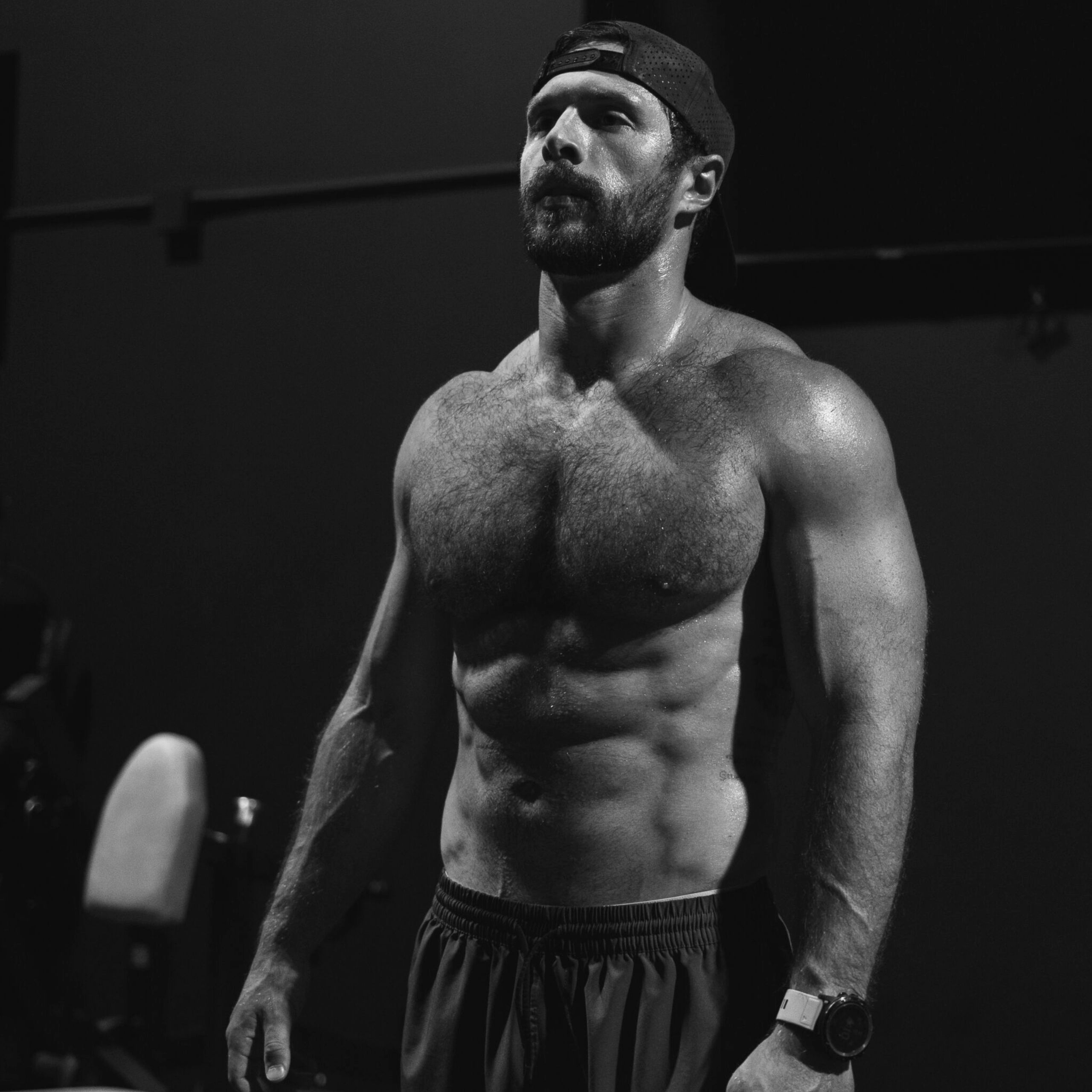 Muscular man with a beard and backward cap standing shirtless in a gym, showing defined chest and abs.