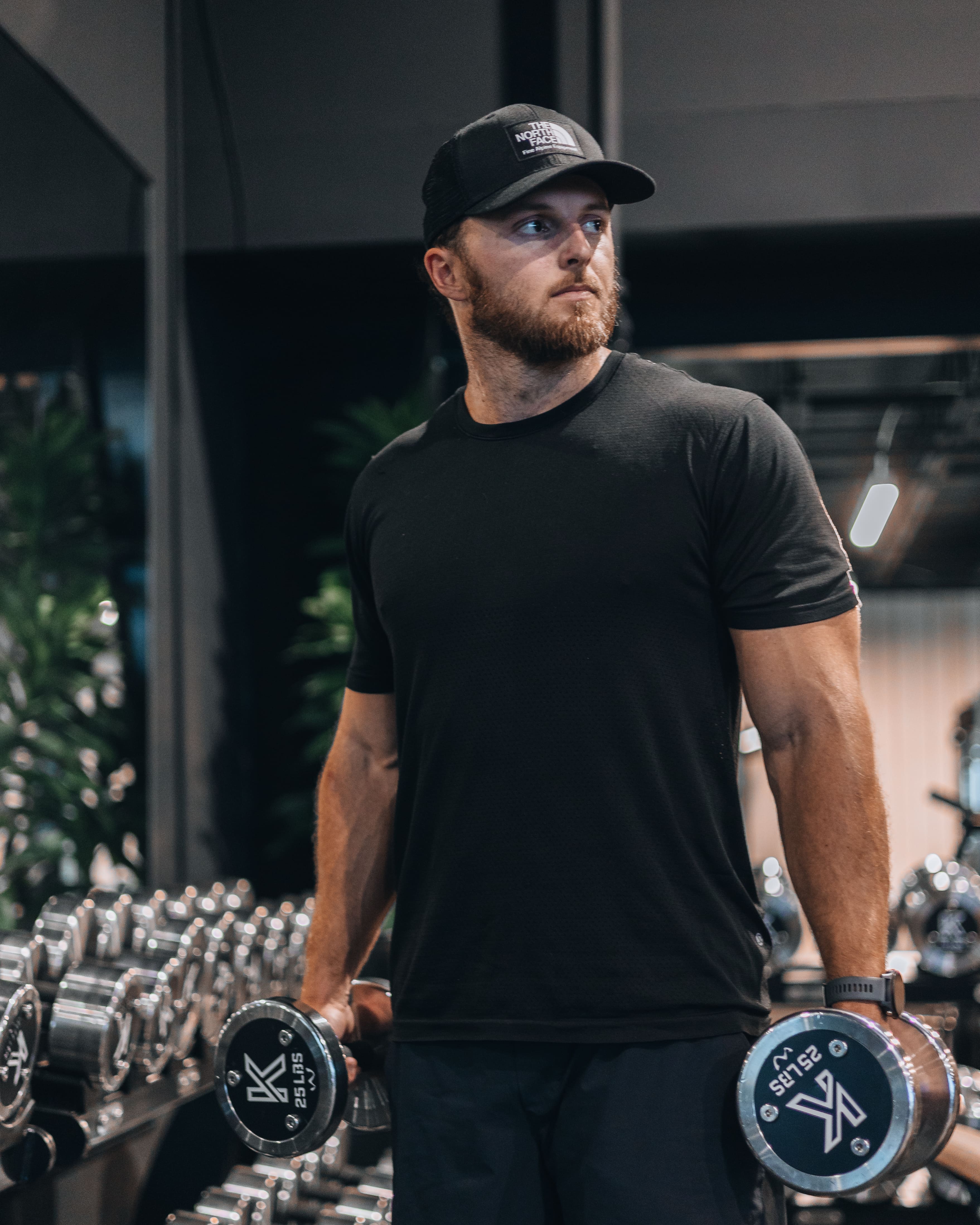 Man in black shirt and cap lifting dumbbells in a gym with mirrors and weights in the background.