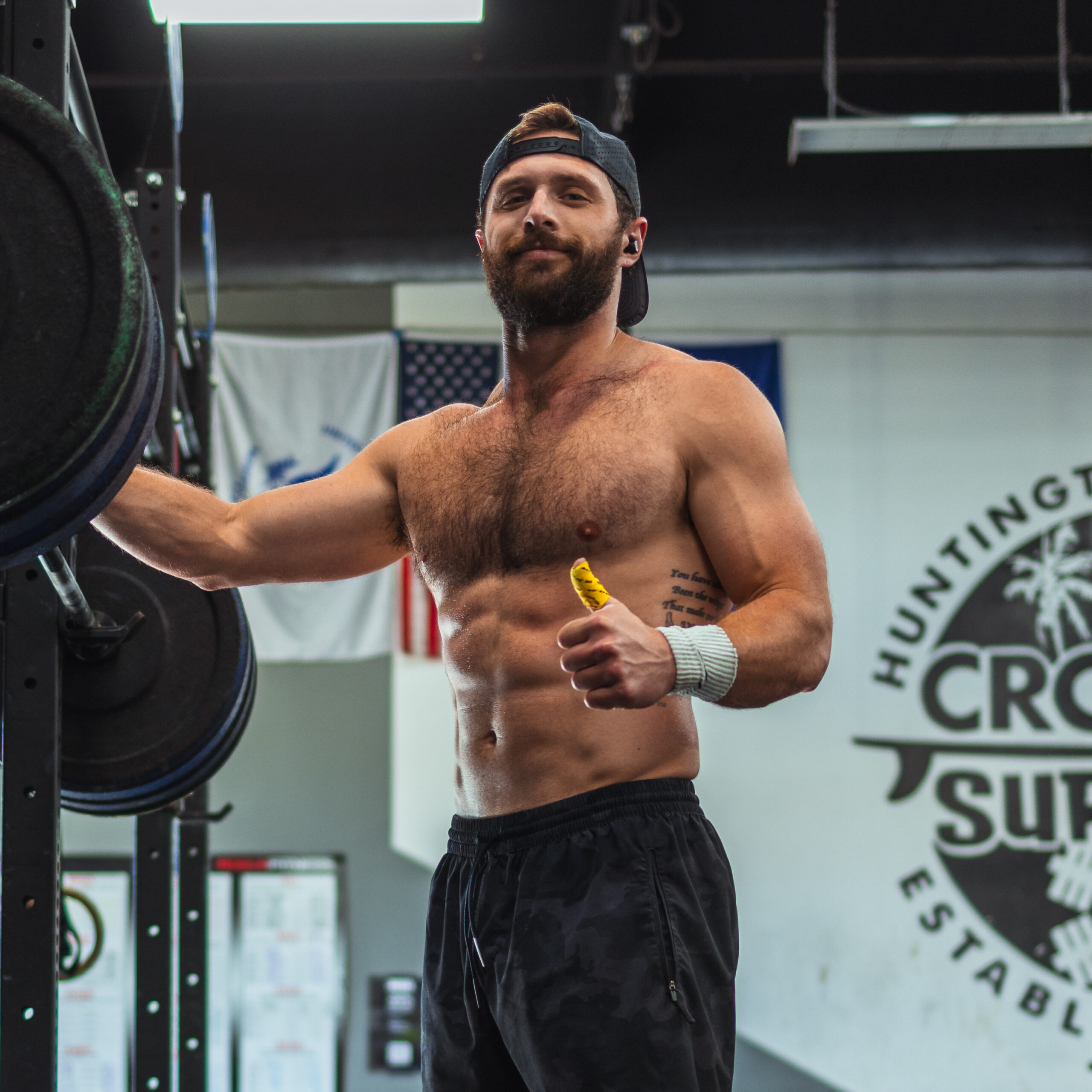 Muscular shirtless man wearing a backward cap and black pants, giving a thumbs-up while standing next to a barbell in a gym.