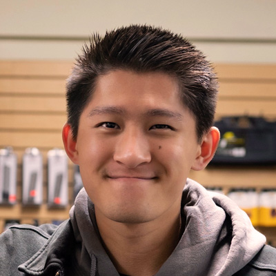 Smiling young man with short dark hair wearing a gray hoodie and dark jacket indoors with shelves and products in the background.