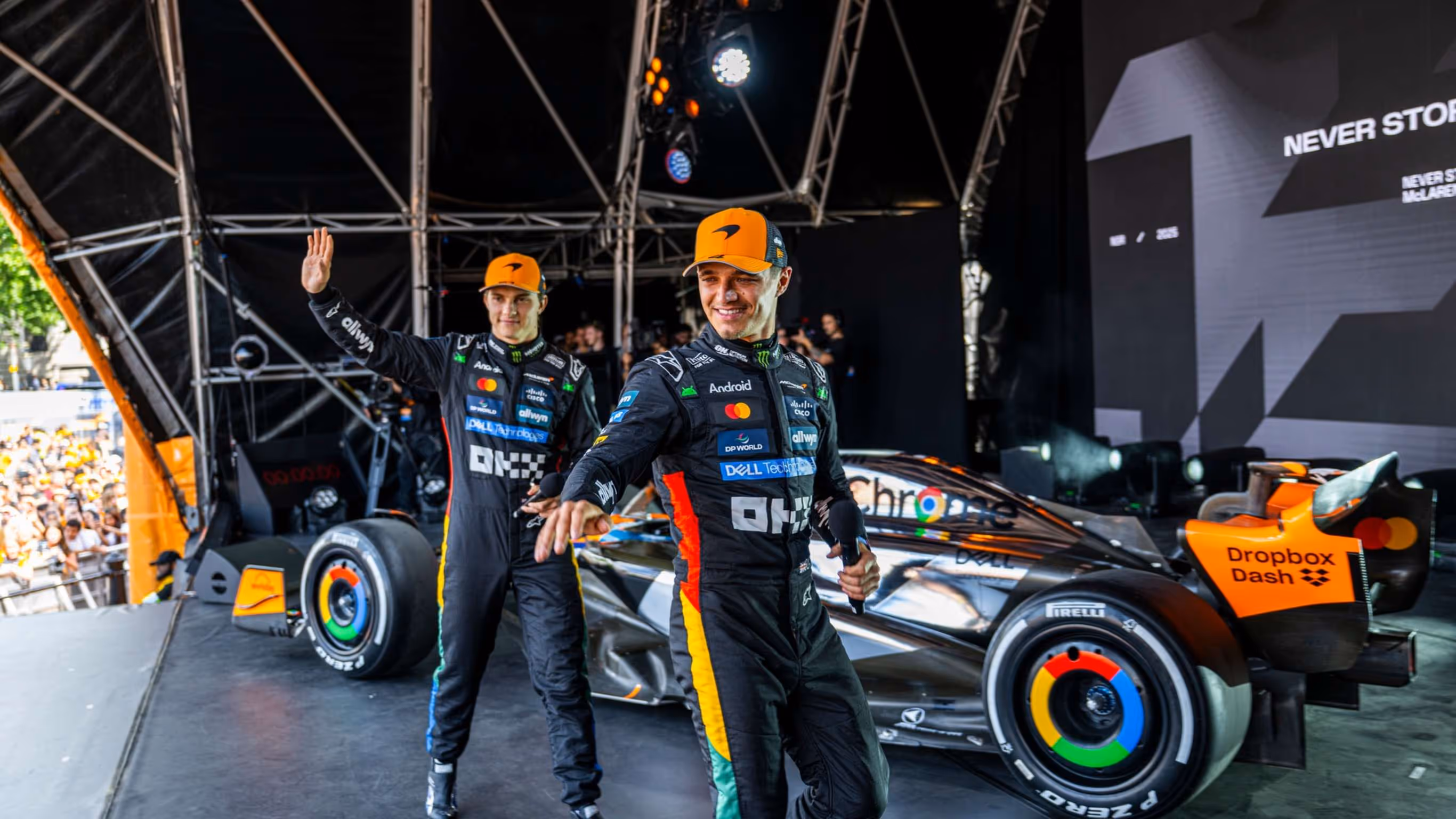 Two race car drivers in black racing suits and orange caps stand next to a silver and orange Formula 1 car on a stage with a crowd in the background.