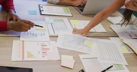 Two people collaborating at a table with charts, graphs, and laptops, discussing printed data sheets.