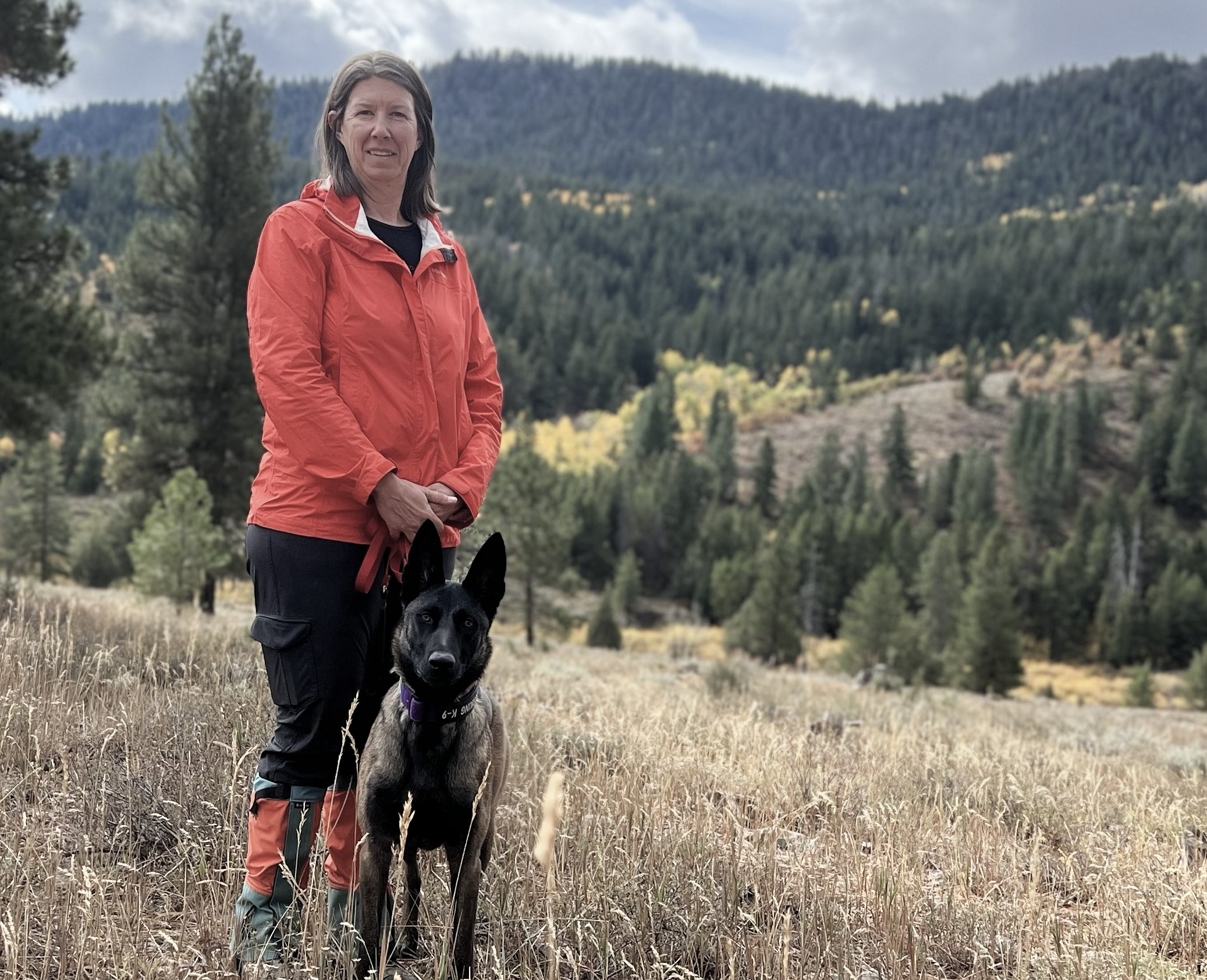 Melissa standing with Cheela, a Belgian Malinois, with mountains in the background