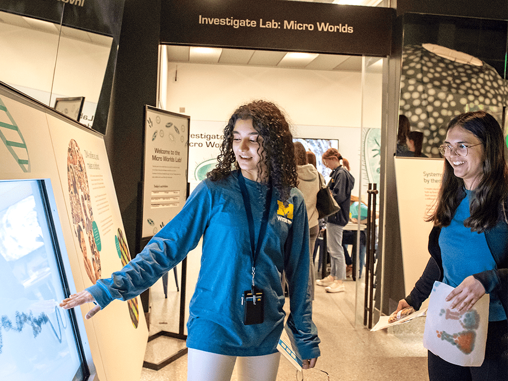 Two students from the Women in Science and Engineering Residence Program (WISE RP) explore the Micro Worlds Investigate Lab interactive exhibit at the University of Michigan Museum of Natural History. They are wearing blue t-shirts with a maize Block M above the acronym WISE-RP. Their faces are lit by the glow of a touchscreen display.