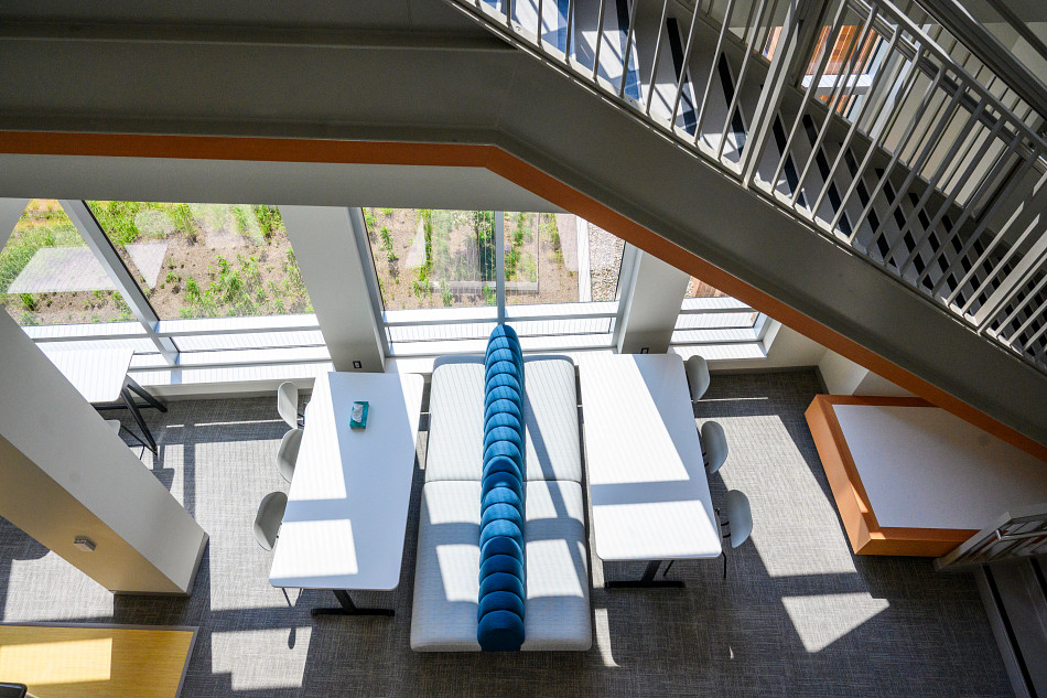 Interior, aerial view of a modern lounge area showing white tables, comfortable benches with a blue backrest, and a staircase, with large windows providing natural light.