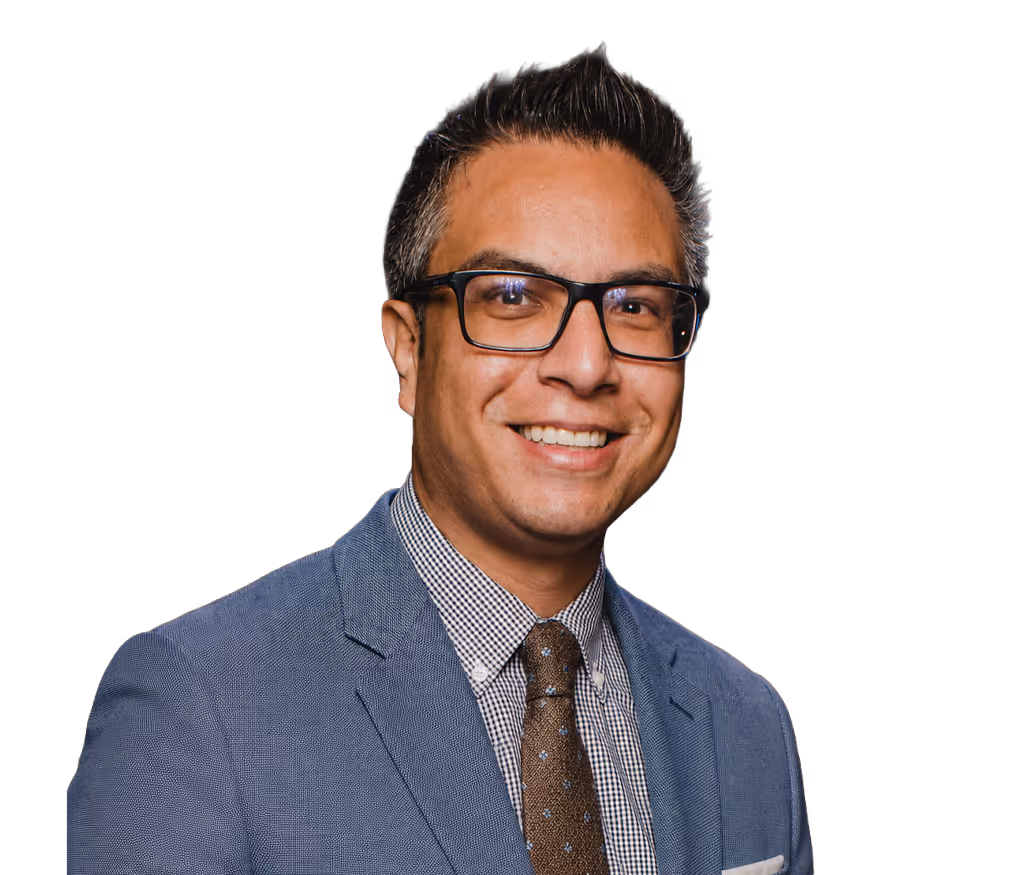 Hombre calvo con gafas redondas oscuras, camisa beige y collar de perlas sonriente.