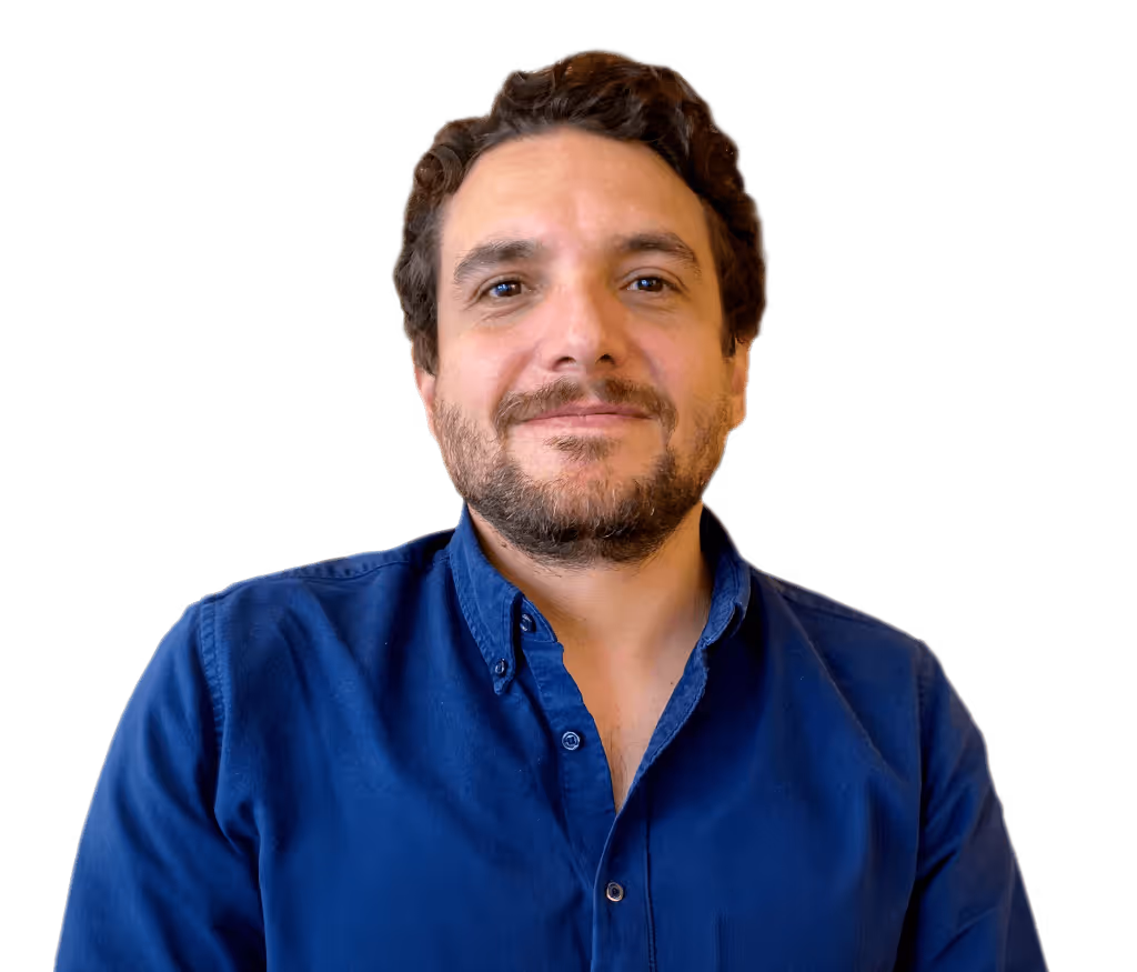 Hombre calvo con gafas redondas oscuras, camisa beige y collar de perlas sonriente.