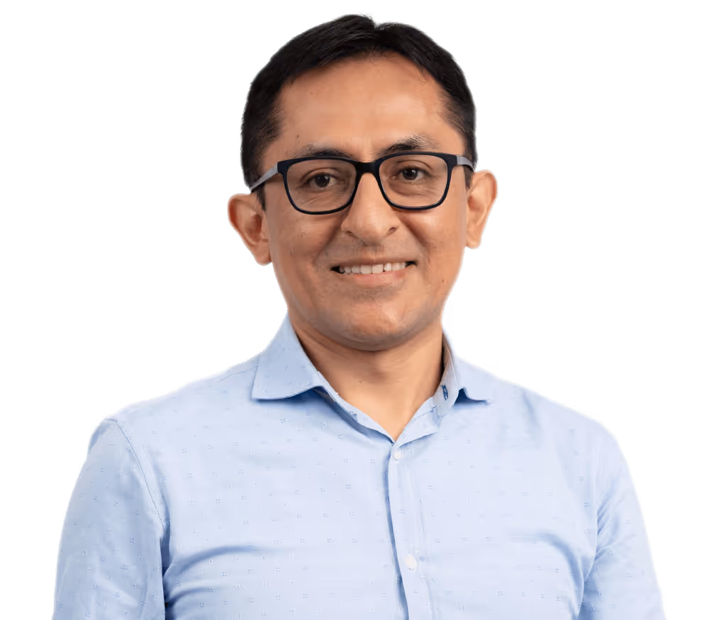 Hombre calvo con gafas redondas oscuras, camisa beige y collar de perlas sonriente.