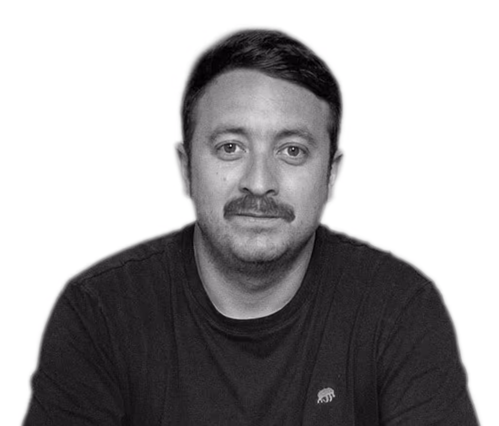 Hombre calvo con gafas redondas oscuras, camisa beige y collar de perlas sonriente.
