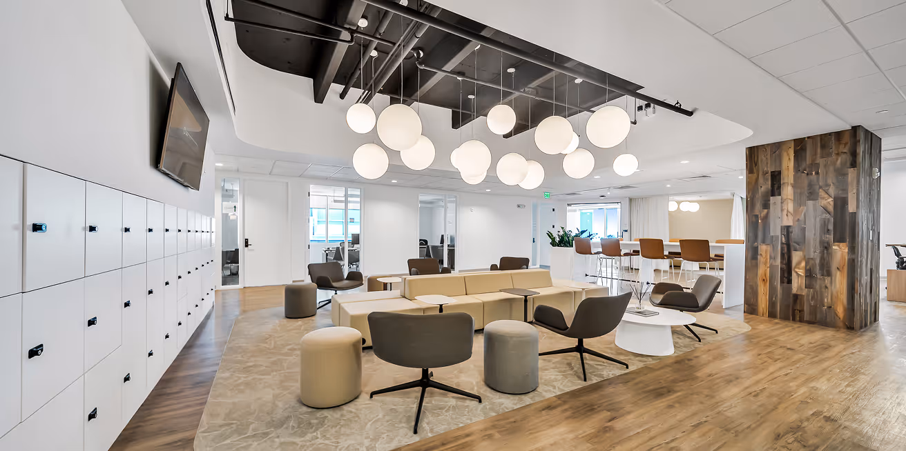 Modern office lounge with beige sectional sofa, gray chairs, round ottomans, wooden floor, hanging globe lights, and white lockers along one wall.