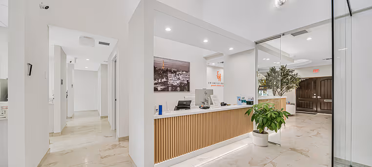Bright modern office reception area with white walls, wooden slatted front desk, potted plants, and marble floor tiles.