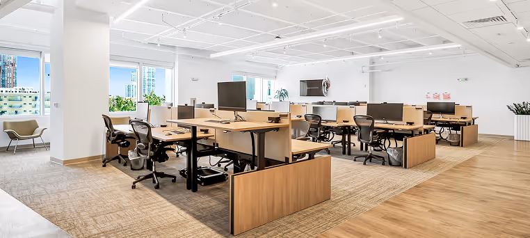 Modern open-plan office with wooden desks, black office chairs, computer monitors, and large windows showing city buildings.