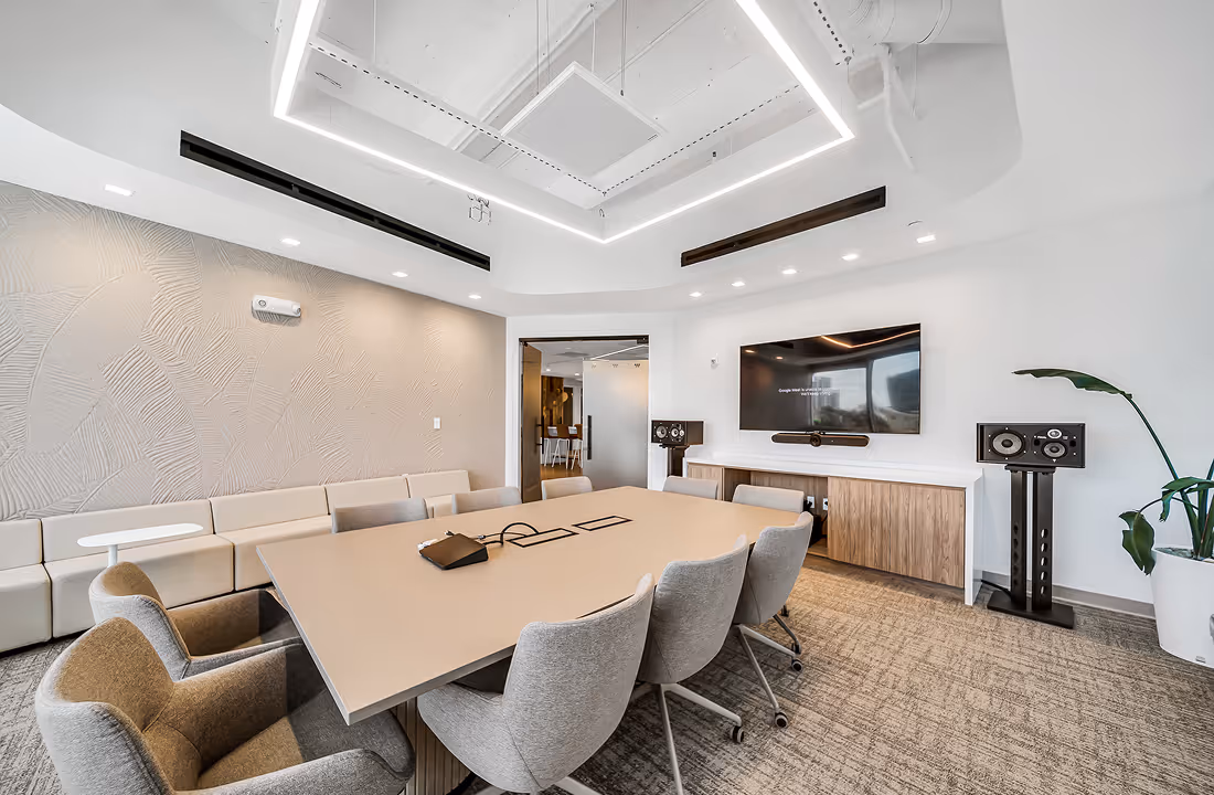 Modern conference room with beige table, gray chairs, wall-mounted TV, speaker system, and corner plant.