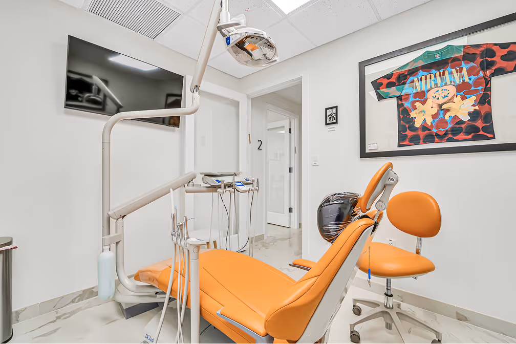 Modern dental office with orange dental chair, dental equipment, a TV on the wall, and framed Nirvana shirt artwork.