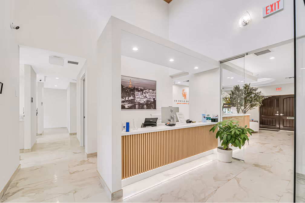Modern healthcare office reception with wooden panel desk, computers, marble floor, and large potted plants near glass doors.