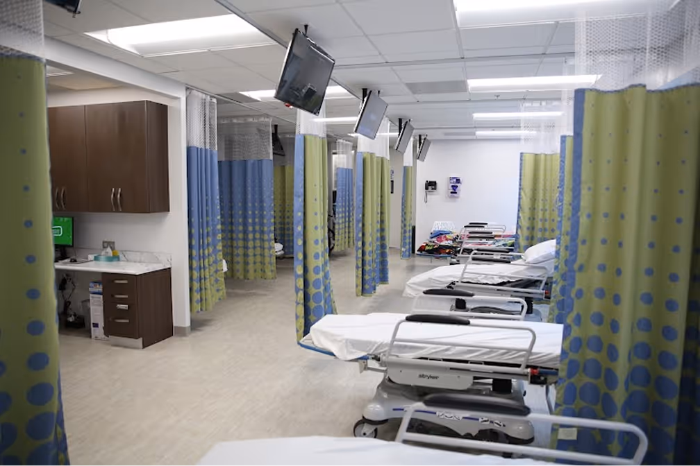 Empty medical examination beds lined up in a row with privacy curtains and monitors attached above each bed in a clinical room.