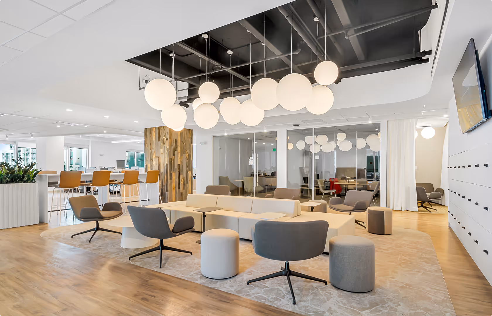 Modern office lounge area with gray and beige chairs, round ottomans, wooden flooring, and spherical pendant lights.