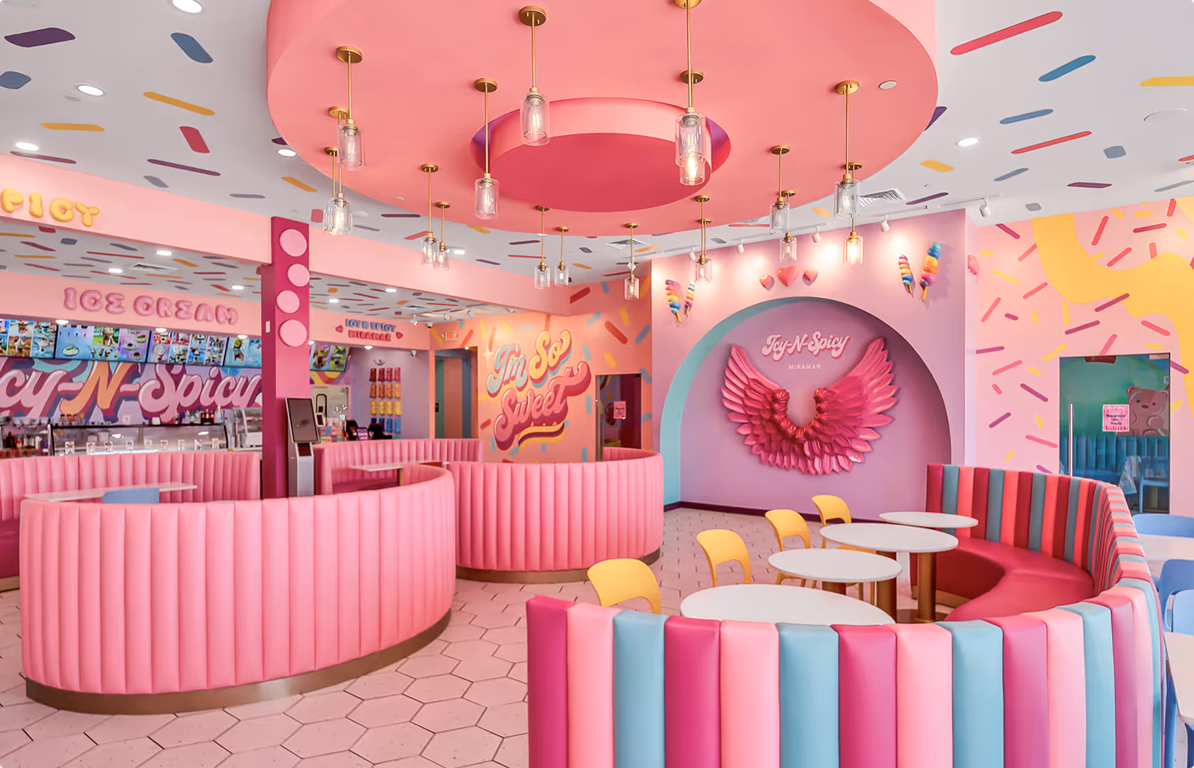 Colorful ice cream shop interior with curved pink booths, round tables, yellow chairs, and wall decoration of pink wings labeled Joy-N-Spicy.