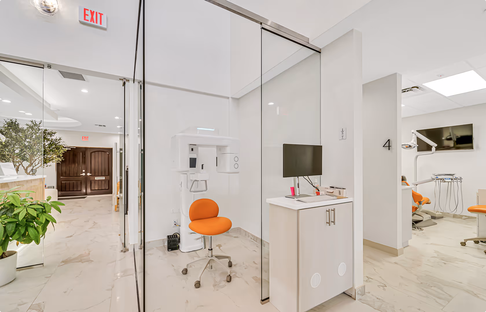 Modern dental office with glass partitions, orange chairs, dental equipment, and a computer workstation.